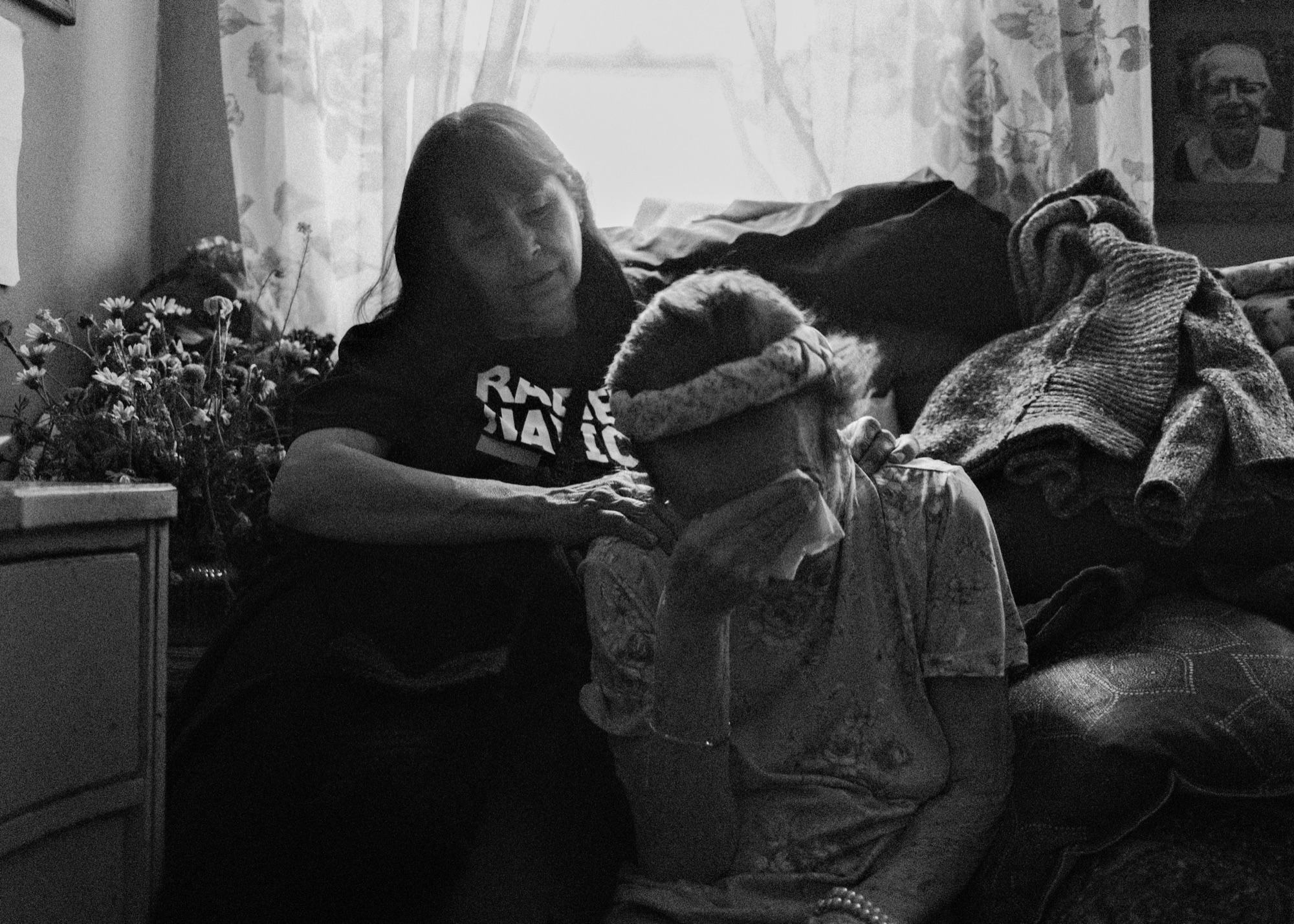 A woman rests her hand on the shoulder of an elderly woman who covers her face, in black and white